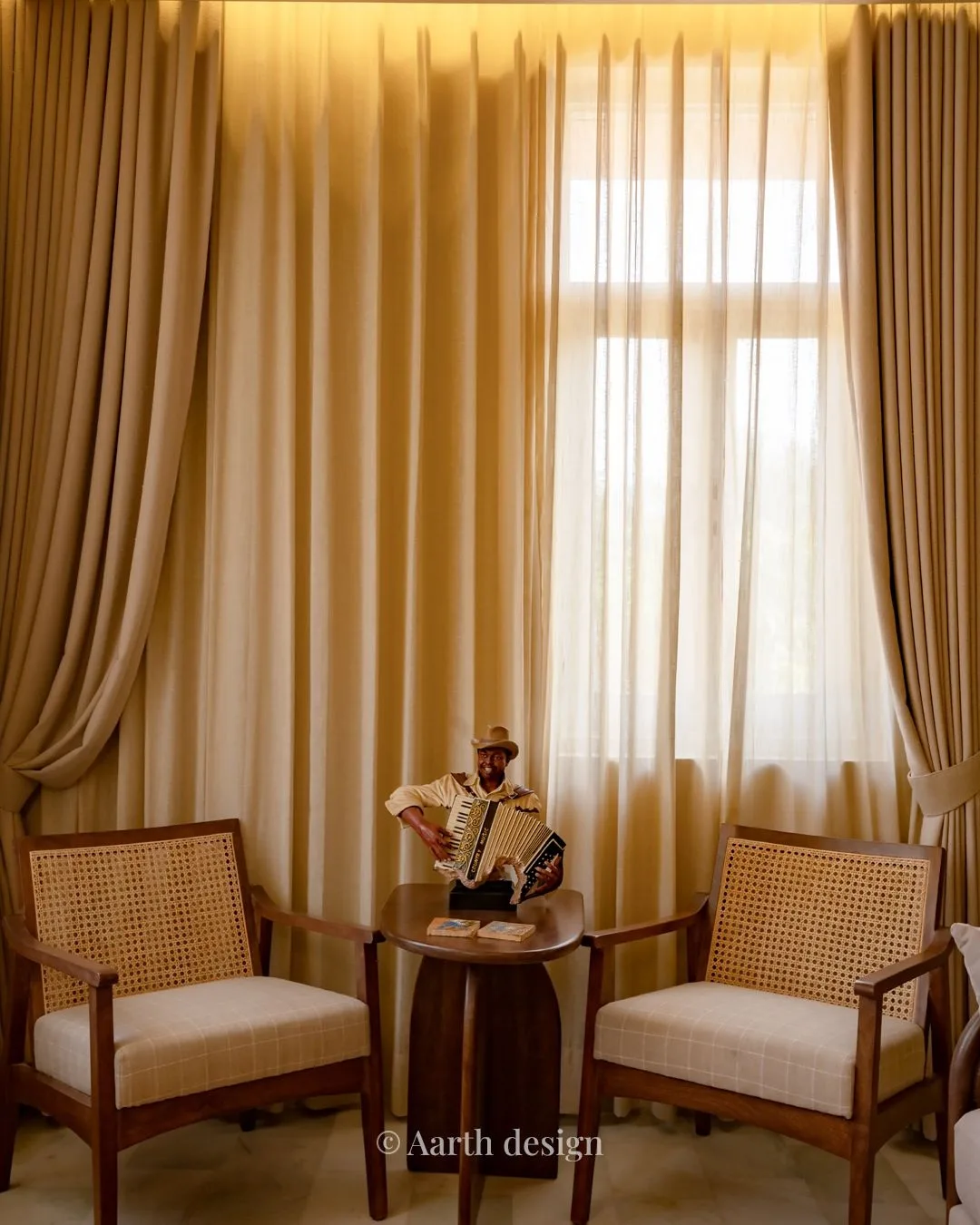 Cane armchairs flanking a round table dressed with folk curios, against sheer curtains