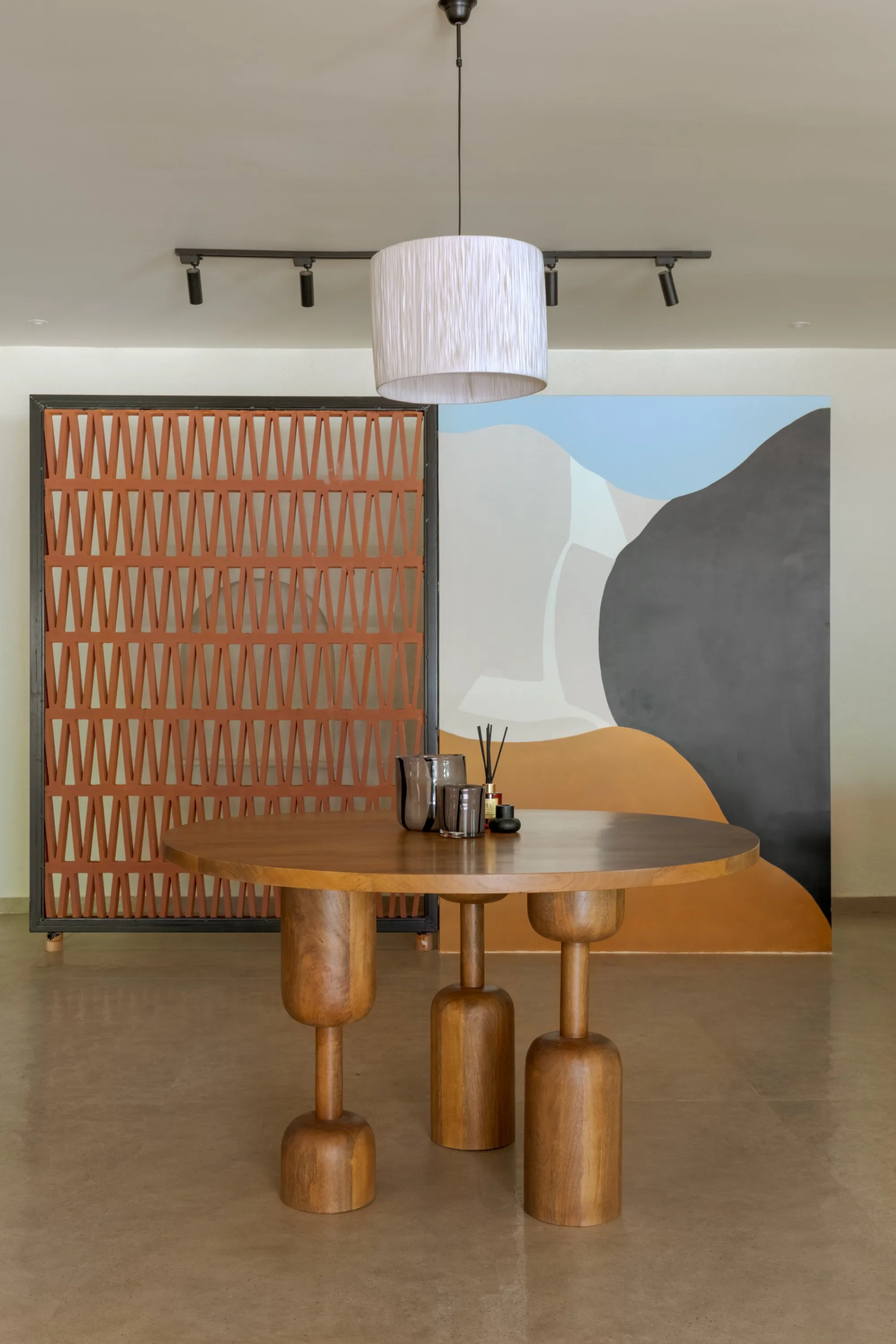 Round dining table on carved wooden legs beneath a drum pendant, with a terracotta jaali screen and a painted wall behind
