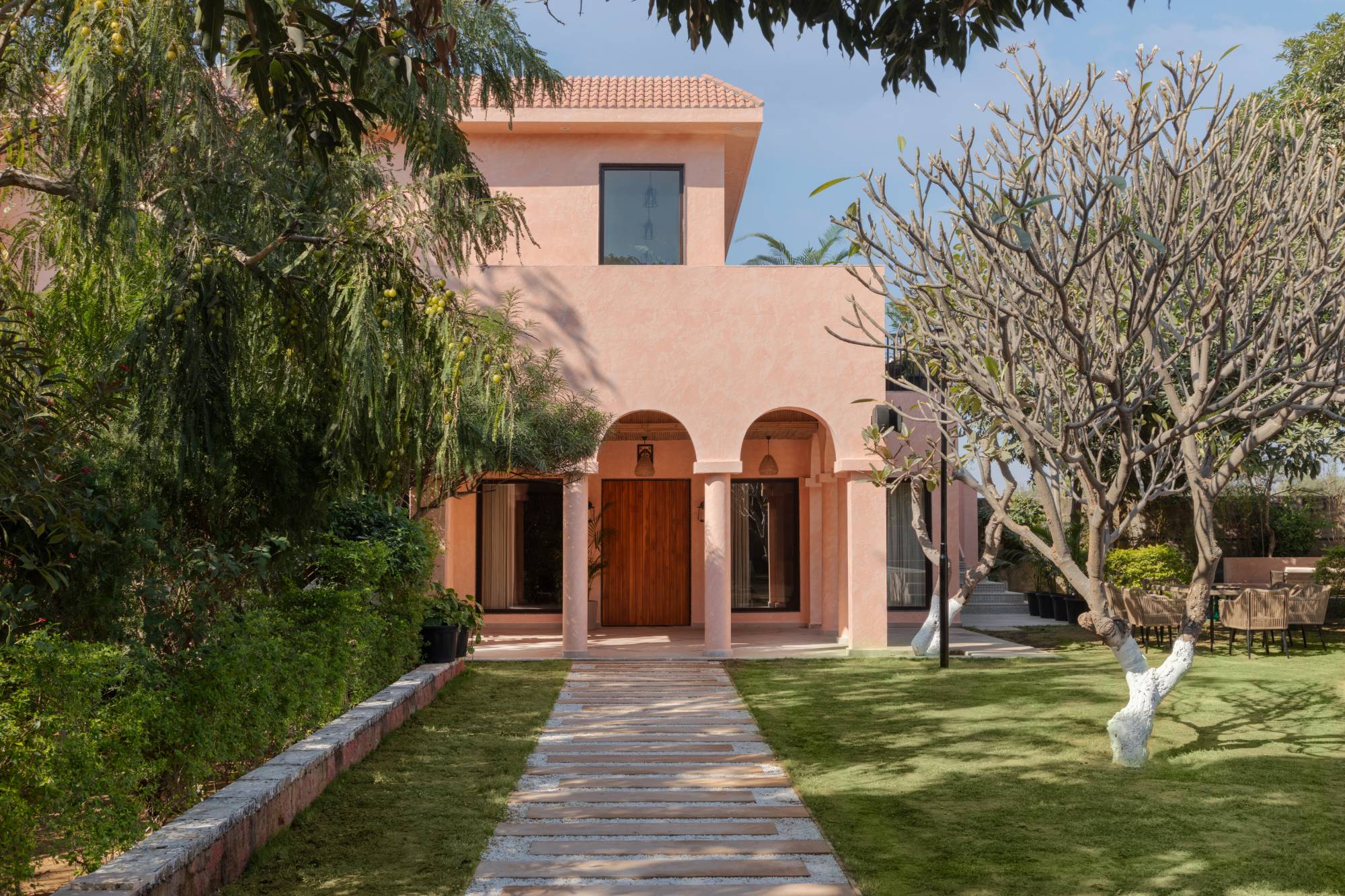 Pink stucco villa with arched portico and a carved wooden door, set behind a lawn and stone pathway