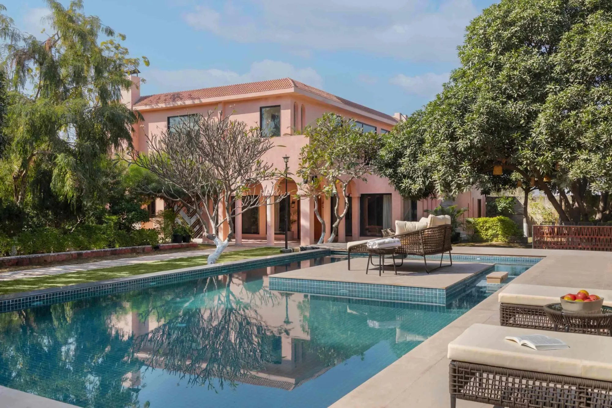 Pink villa seen across a turquoise pool, with frangipani trees and woven loungers on the terrace